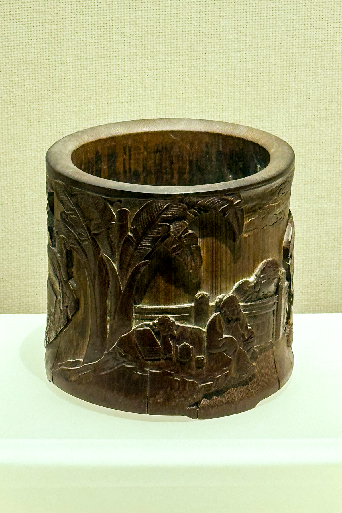 A dark brown carved wooden cylindrical brush holder shows a landscape scene, reflecting artistic traditions relevant to.
